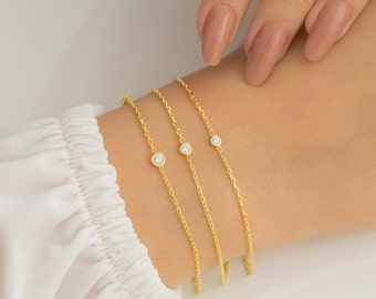 May include: Three gold chain bracelets with a single small clear gemstone on each. The bracelets are layered on a person's wrist.