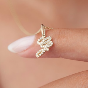Könnte beinhalten: Eine goldene Halskette mit einem diamantbesetzten Buchstabenanhänger. Der Anhänger ist in Form des Buchstabens "G" gestaltet und wird von einem Finger gehalten.