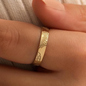May include: A gold-colored ring with floral engravings, including a sunflower design. The ring is worn on a finger, showcasing the detailed craftsmanship of the jewelry. The ring is a piece of fashion jewelry.
