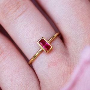 May include: A gold ring with a rectangular red gemstone. The ring is on a person's finger.