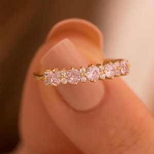 Könnte beinhalten: Ein Goldring mit einer Reihe von rosafarbenen Edelsteinen. Der Ring ist mit kleinen, runden Steinen besetzt.