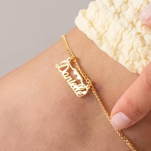 May include: A gold-coloured anklet with the name "Danielle" in a cursive font. The anklet features a delicate chain and a charm with the name. The anklet is worn on an ankle.