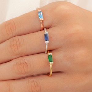 May include: Three gold rings with different colored gemstones. The top ring has a light blue gemstone, the middle ring has a dark blue gemstone, and the bottom ring has a green gemstone. Each ring has small clear gemstones on either side of the main gemstone.