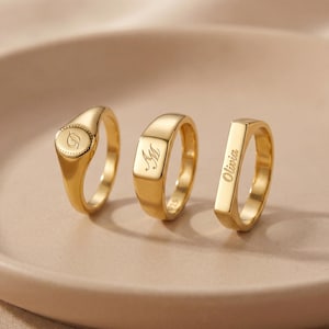 May include: Three gold signet rings on a neutral surface. The rings vary in shape: one oval, one rectangular, and one with a flat top. Each ring has engraved details, including initials and the name "Olivia".