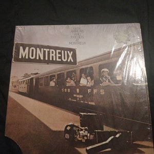 Op de afbeelding: Een vintage vinyl platenhoes met een zwart-witfoto van een trein met de tekst "MONTREUX" op een bord erboven. Het album is getiteld "Gene Ammons and Friends at Montreux".