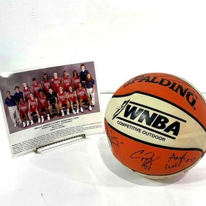 Puede incluir: Balón de baloncesto Spalding WNBA naranja y blanco con texto negro y firmas de jugadores. También hay una fotografía enmarcada del equipo olímpico femenino de baloncesto de EE. UU. de 2004.