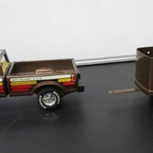 Op de afbeelding: Een bruine speelgoed pick-up truck met een bijpassende bruine aanhanger. De truck heeft een witte en oranje streep langs de zijkant en de woorden "FLYNN'S HORSE RANCH" staan ​​op de zijkant gedrukt. De aanhanger heeft de woorden "FLYNN'S HORSE RANCH" op de zijkant gedrukt.