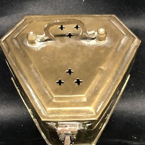 May include: A vintage brass box with a geometric design. The box has a handle, decorative cross-shaped cutouts, and a latch closure. The brass has a warm, aged patina, suggesting its age. The box is likely used for storage or display.