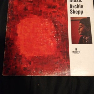 Könnte beinhalten: Ein Schallplattencover mit einem abstrakten roten und schwarzen Gemälde auf der linken Seite. Die rechte Seite zeigt den Text "Fire Music Archie Shepp" und ein Schwarzweißfoto eines Mannes mit einem schwarzen Hut. In der rechten unteren Ecke steht der Text "Impulse!" und "Stereo A-96".