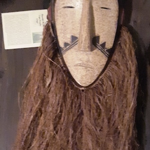 Puede incluir: Una máscara de madera con la parte superior negra y la cara de color marrón claro. La máscara tiene una nariz larga y fina y ojos pequeños y cerrados. La máscara está decorada con líneas negras y una barba marrón hecha de fibras.