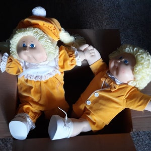 May include: Two Cabbage Patch Kids dolls in a cardboard box. Both dolls are wearing yellow outfits with white accents. The dolls have blonde yarn hair and blue eyes.