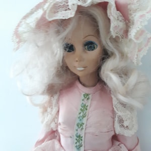 May include: A vintage porcelain doll with blonde hair, blue eyes, and a pink dress with lace trim. The doll is wearing a pink bonnet with lace trim.