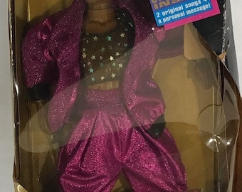 May include: A pink and black doll of MC Hammer wearing a pink jacket, a black and gold star-patterned shirt, and pink pants. The doll comes with a real cassette tape inside the box. The box has a gold background with the text "REAL CASSETTE INSIDE" and "2 original songs + a personal message!"