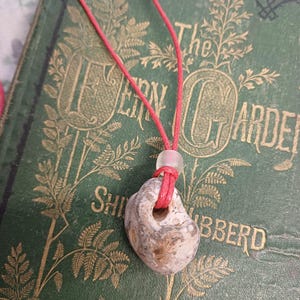 May include: A red cord necklace with a grey and white stone pendant. The stone has a hole in the centre and is tied to the cord with a white bead.
