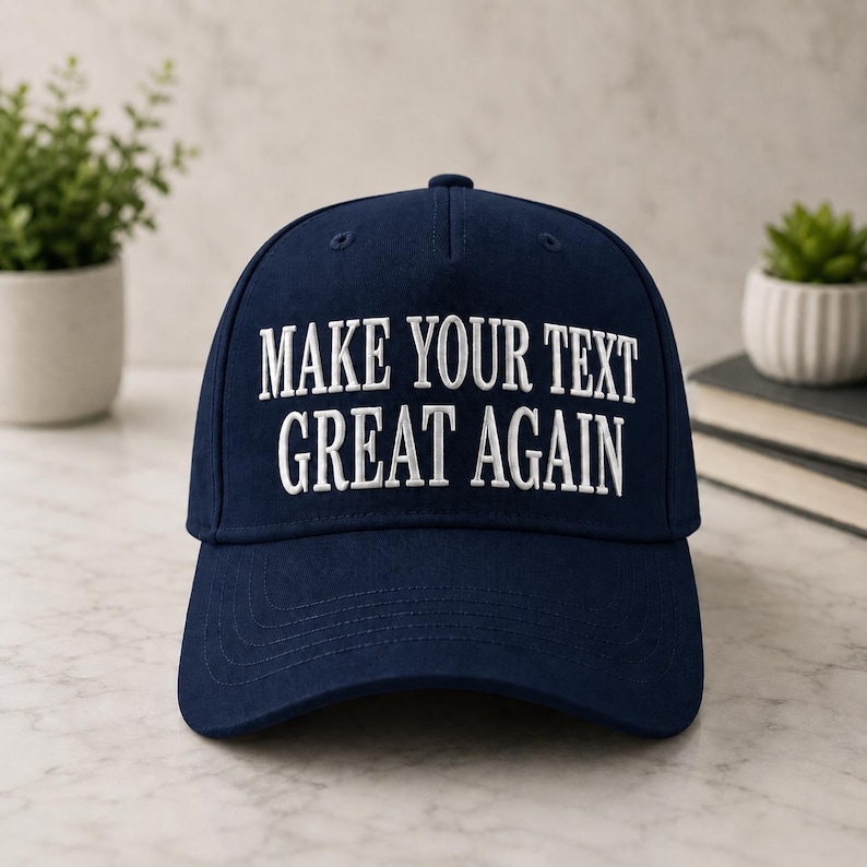 Może przedstawiać: Granatowa czapka bejsbolowa z białym haftem &bdquo;MAKE YOUR TEXT GREAT AGAIN&rdquo;. Czapka jest prezentowana na marmurowej powierzchni, z małymi roślinami doniczkowymi w tle.