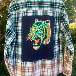 Puede incluir: Una camisa de franela a cuadros verde y blanca con un parche gráfico de tigre en la espalda. La mitad inferior de la camisa es un patrón de cuadros diferente con marrón, blanco y verde.