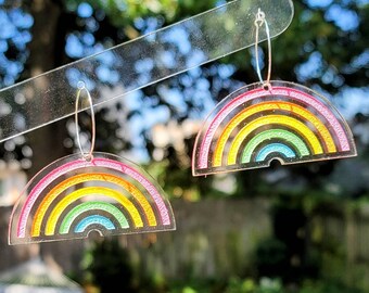 May include: A pair of rainbow earrings made of clear acrylic with a silver hoop. The earrings are shaped like rainbows with multiple colors, including pink, yellow, green, blue, and purple.