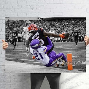 May include: A black and white photograph of a football game in action. A player in a black jersey with the name "Higgins" on the back is reaching for the ball while another player in a purple jersey with the name "Evans" is falling. The image is set in a stadium.