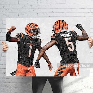 May include: Watercolor-style print of two football players in black and orange uniforms, helmets, and pads. One player has the number 1, the other has the number 5. The background is a white and gray watercolor effect.