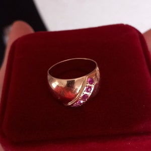 Op de afbeelding: Een gouden ring met een diagonale rij roze edelstenen die in de band zijn gezet. De ring wordt gepresenteerd in een rode fluwelen ringdoos.