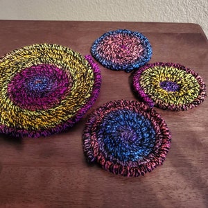 Puede incluir: Cuatro posavasos hechos a mano con un diseño en espiral. Los posavasos presentan una vibrante paleta de colores de amarillo, morado, azul y rosa. Los posavasos están hechos de un material tejido y están dispuestos sobre una superficie de madera oscura.