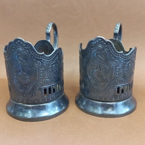 Peut inclure: Deux porte-gobelets en métal argenté avec des motifs ornés et des poignées. Les porte-gobelets sont sur une surface brune.