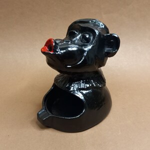 Puede incluir: Cenicero de cerámica negro con forma de mono con la boca pintada de rojo. El mono tiene un pequeño cuenco para ceniza en la parte posterior de su cabeza.