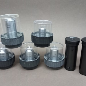 May include: A set of six microscope objectives, including four objective lenses in clear plastic containers and two black metal objective lenses. The objective lenses have various magnification levels, including 32x, 100x, 40x, and 160x.