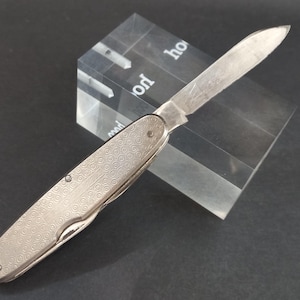 May include: A silver pocket knife with an open blade, set against a clear, rectangular block. The knife's handle features a detailed, circular pattern. The blade is extended, showcasing its sharp edge. The background is a dark surface.