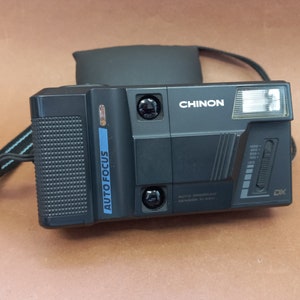 May include: A black and silver Chinon Auto Focus DX camera with a built-in flash. The camera has a blue "Auto Focus" label on the side.
