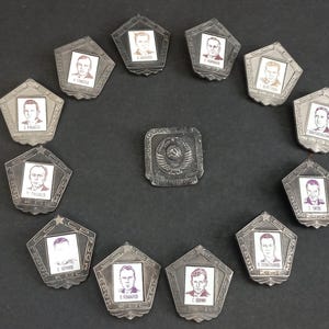 Op de afbeelding: Een verzameling vintage Sovjet-tijdperkspelden en -badges. De spelden zijn zilverkleurig, vijfhoekig en tonen portretten van mannen. Een centrale badge toont een hamer en sikkel embleem. Op een donkere ondergrond.