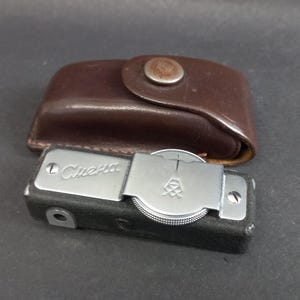 Puede incluir: Una cámara vintage plateada y negra con el nombre "Cuera" grabado en la parte superior. La cámara va acompañada de una funda de cuero marrón con un cierre metálico. La cámara tiene un dial y una lente.