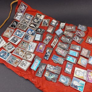 May include: A collection of vintage metal pins with various designs, including portraits and space-themed imagery, displayed on a red fabric. The pins have silver-colored frames and are arranged in a scattered pattern.