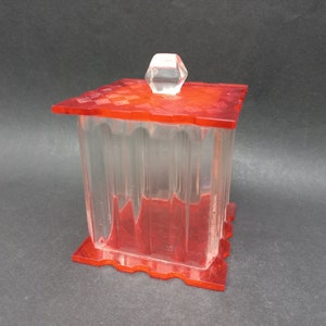 Puede incluir: Una caja de plástico transparente con una tapa roja y un pomo de plástico transparente en la parte superior. La caja tiene un borde decorativo festoneado.