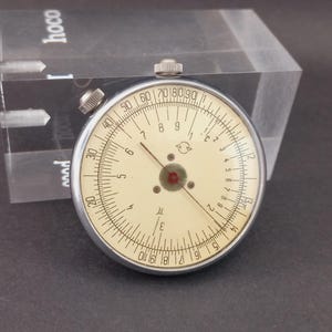 May include: A vintage circular slide rule with a cream face, black markings, and a red centre. The slide rule is in a silver metal frame. A clear acrylic block is in the background.