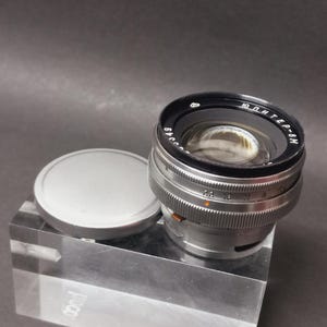 Può includere: Un obiettivo per fotocamera argento e nero con un copriobiettivo. L'obiettivo ha un anello nero con testo bianco e un corpo argento con segni di misurazione. Il copriobiettivo è rotondo e argentato.