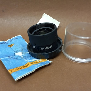 Puede incluir: Un objetivo de teleconvertidor negro con el texto "TK-2M 072387" impreso en el lateral. El objetivo está en una caja de plástico transparente.