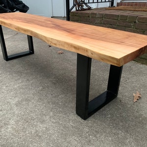 May include: A wooden bench with a live edge top and black metal legs. The bench is made of natural wood and has a rustic look.