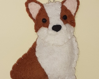 May include: A felt corgi dog ornament with a silver chain for hanging. The dog is brown and white, with black eyes and a black nose. The ears are brown with pink inner ears. The ornament is handmade and has a stitched outline.