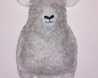 May include: A handmade grey felt sheep decoration with white details. The sheep has a fluffy body, white ears, and white feet. A silver string is attached for hanging. The sheep's face features a white muzzle and a black stitched mouth.