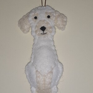 May include: A handmade white felt dog ornament with tan accents on the ears and face. The dog has black eyes and a black nose. A brown string is attached for hanging. The ornament is a whimsical decoration.