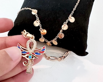 Könnte beinhalten: Goldfarbene Halskette mit einem Anhänger in Form eines Ankh, mit roten, blauen und weißen Emailleflügeln und einem türkisfarbenen Stein. Eine zweite Halskette mit kleinen goldenen Scheiben ist ebenfalls vorhanden. Der Schmuck wird auf einer schwarzen Oberfläche präsentiert.