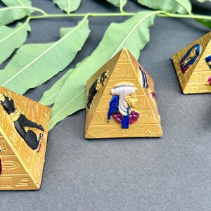 Pode incluir: Três objetos decorativos dourados em forma de pirâmide com desenhos egípcios. Cada pirâmide apresenta hieróglifos e detalhes coloridos em relevo de faraós e outras figuras. Folhas verdes no fundo.
