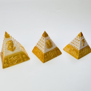 Può includere: Tre scatole decorative a forma di piramide con dettagli dorati e crema. Ogni piramide presenta disegni in rilievo, tra cui geroglifici e motivi egizi. Le piramidi variano leggermente nelle dimensioni e sono disposte su una superficie bianca.