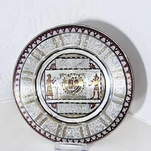 May include: Decorative silver plate with an Egyptian theme. The plate features intricate engravings, including hieroglyphs and figures, with gold and burgundy accents. The rim has a burgundy border with a repeating pattern.