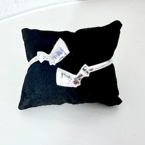 Puede incluir: Un brazalete plateado con un diseño ornamentado, exhibido sobre una superficie de terciopelo negro. El brazalete presenta un motivo detallado, posiblemente de inspiración egipcia. El diseño es visible en la parte delantera del brazalete.