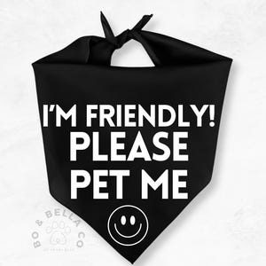 Op de afbeelding: Zwarte hondenbandana met witte tekst "I'M FRIENDLY! PLEASE PET ME" en een smiley. De bandana is bovenaan vastgeknoopt. De linker benedenhoek heeft de tekst "BO & BELLA CO all about pets".