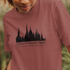May include: A dusty rose t-shirt featuring a black silhouette of a mountain range with the text "VISTA HIGHLINE TRAIL" and "EST. 2014". The shirt is made of a soft material and has short sleeves.