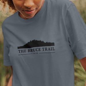 Könnte beinhalten: Ein schieferblaues T-Shirt mit dem Text "THE BRUCE TRAIL" und einer Waldsilhouette. Das Shirt besteht aus einem weichen Material und wird im Freien getragen.