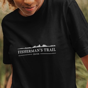 Könnte beinhalten: Schwarzes T-Shirt mit dem weißen Text "Fisherman's Trail" und einer Bergketten-Grafik. Das T-Shirt wird von einer Person in einer natürlichen Umgebung im Freien getragen, mit grünem Laub im Hintergrund.
